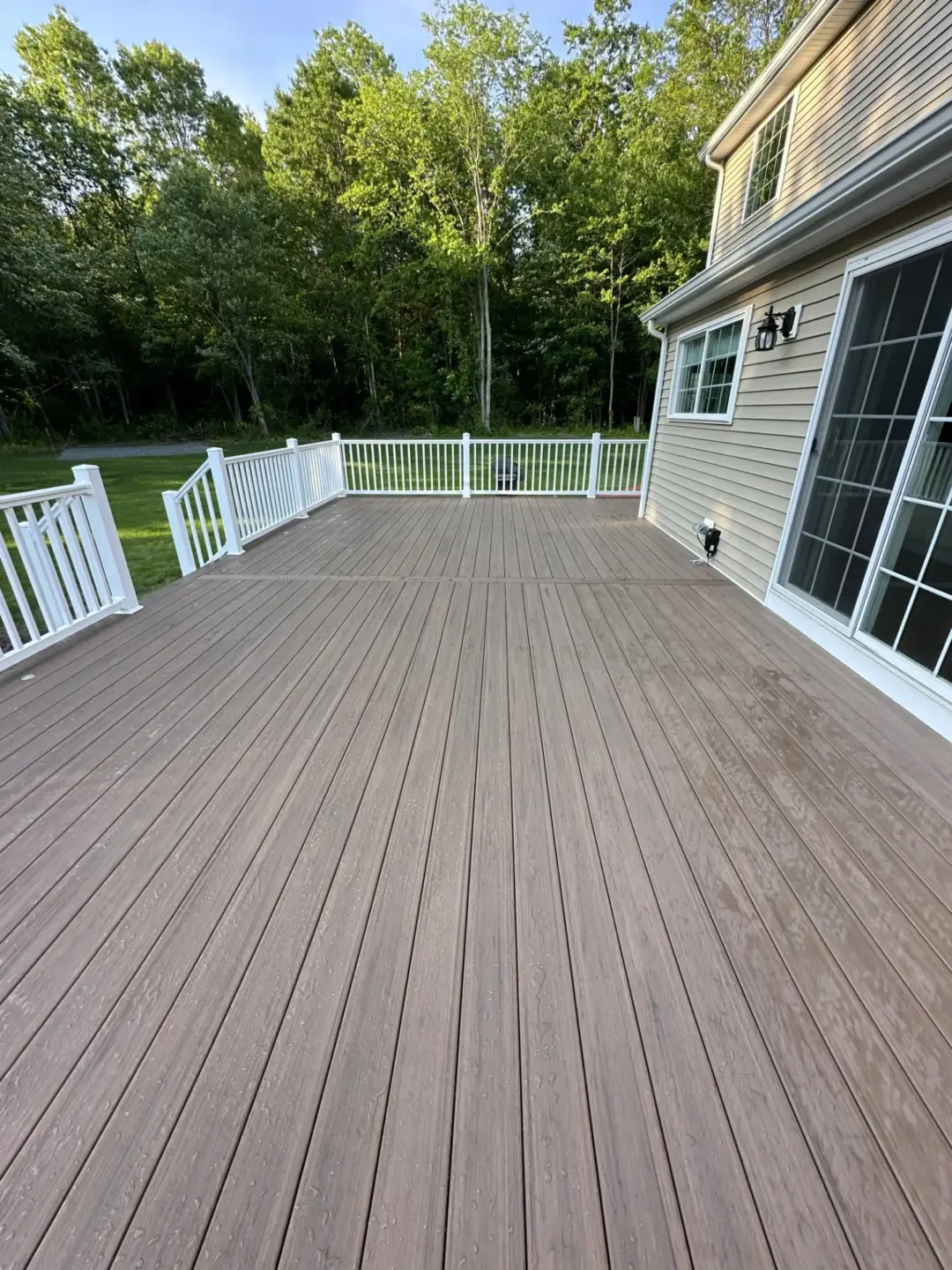 Spacious composite deck with white railing adjacent to house and wooded backyard